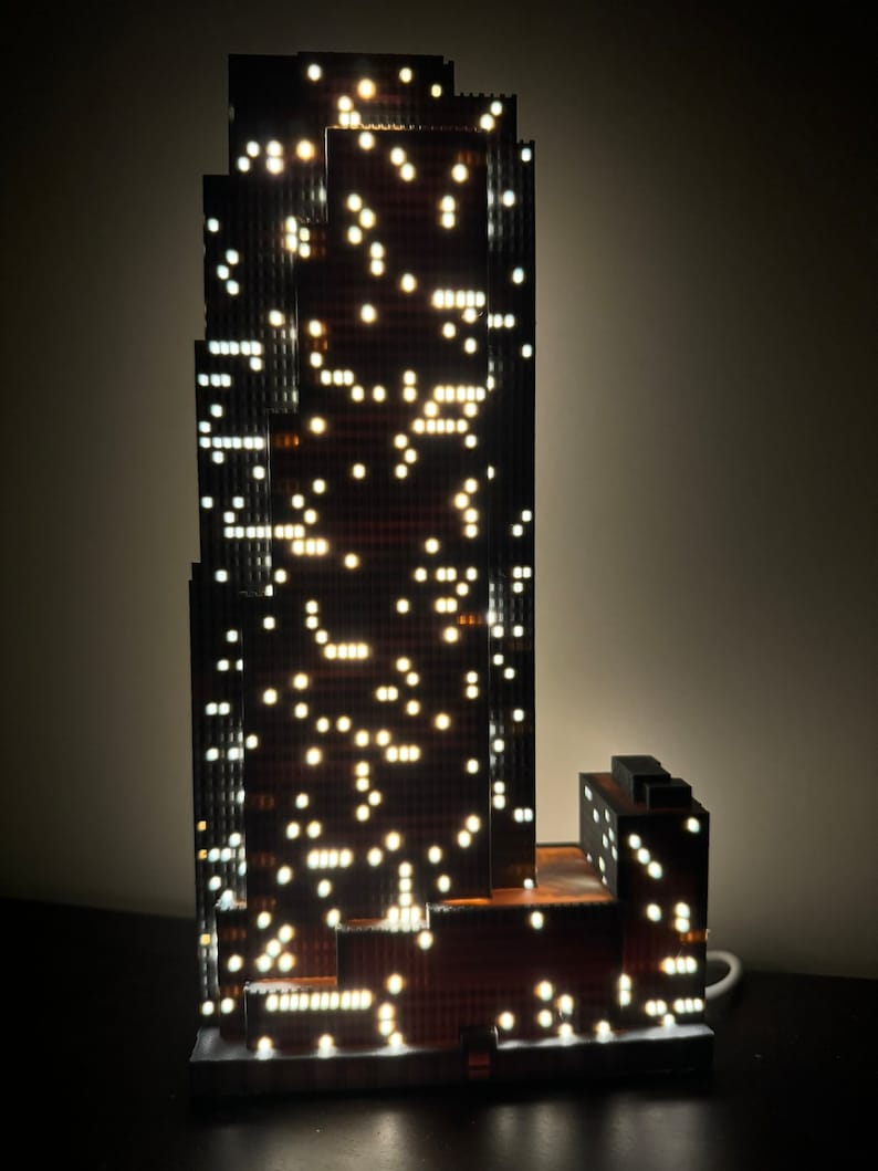 May include: A black and brown skyscraper model with white lights illuminating the windows. The model is lit from within and has a cord attached to the base.