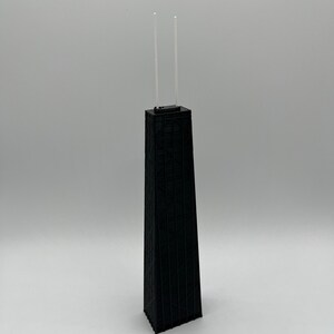 May include: A black architectural model of a skyscraper with two thin, clear antennas extending from the top. The building has a detailed, textured facade and stands on a white surface.