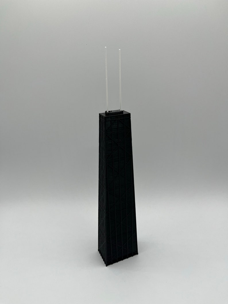 May include: A black architectural model of a skyscraper with two thin, clear antennas extending from the top. The building has a detailed, textured facade and stands on a white surface.