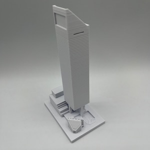 May include: A white 3D printed model of a skyscraper with a small building at the base.