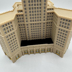 May include: A beige architectural model of a building with multiple stories and numerous windows. The model has a detailed facade with rectangular windows and decorative elements. The building's design includes a central tower and lower wings, with a dark interior space.