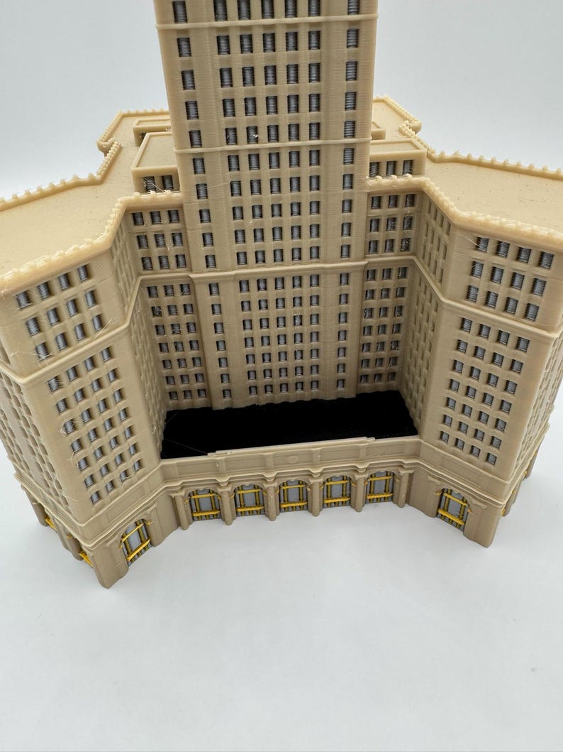May include: A beige architectural model of a building with multiple stories and numerous windows. The model has a detailed facade with rectangular windows and decorative elements. The building's design includes a central tower and lower wings, with a dark interior space.