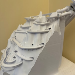 May include: White 3D printed model of a mountain with a winding track and a tower at the top.