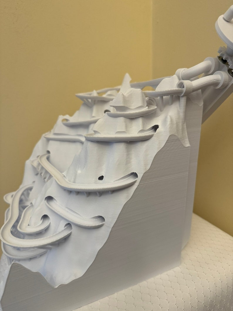 May include: White 3D printed model of a mountain with a winding track and a tower at the top.