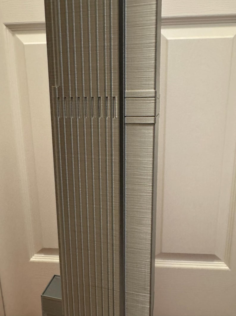 K&ouml;nnte beinhalten: Detailliertes Modell eines Wolkenkratzers mit strukturierter, metallischer Oberfl&auml;che. Die Fassade des Geb&auml;udes weist vertikale Linien und vertiefte Abschnitte mit rechteckiger Basis auf. Das Modell steht vor einem neutralen Hintergrund.