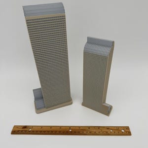 May include: Two miniature beige and gray skyscraper models with detailed window patterns. A wooden ruler with inch markings is placed in front of the models. The buildings have a rectangular shape with a textured facade.