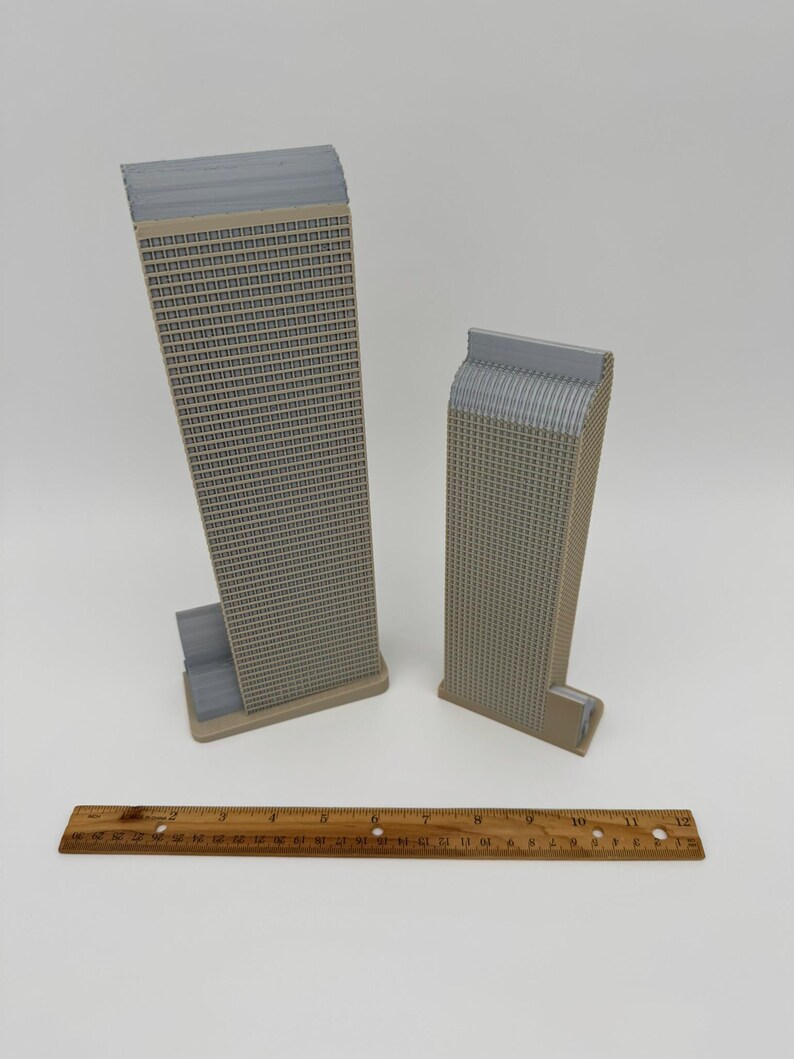 May include: Two miniature beige and gray skyscraper models with detailed window patterns. A wooden ruler with inch markings is placed in front of the models. The buildings have a rectangular shape with a textured facade.