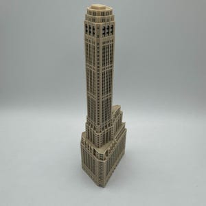 Op de afbeelding: Een gedetailleerd miniatuurmodel van een hoge, beige wolkenkrabber met veel ramen. Het model is gemaakt van plastic en heeft een licht getextureerd oppervlak.