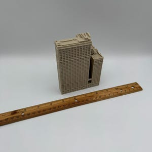 May include: A beige miniature model of a building with a rectangular shape and multiple windows. A wooden ruler is placed diagonally across the image, measuring up to 12 inches. The building model has a detailed facade and a flat roof.