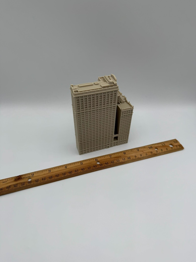 May include: A beige miniature model of a building with a rectangular shape and multiple windows. A wooden ruler is placed diagonally across the image, measuring up to 12 inches. The building model has a detailed facade and a flat roof.