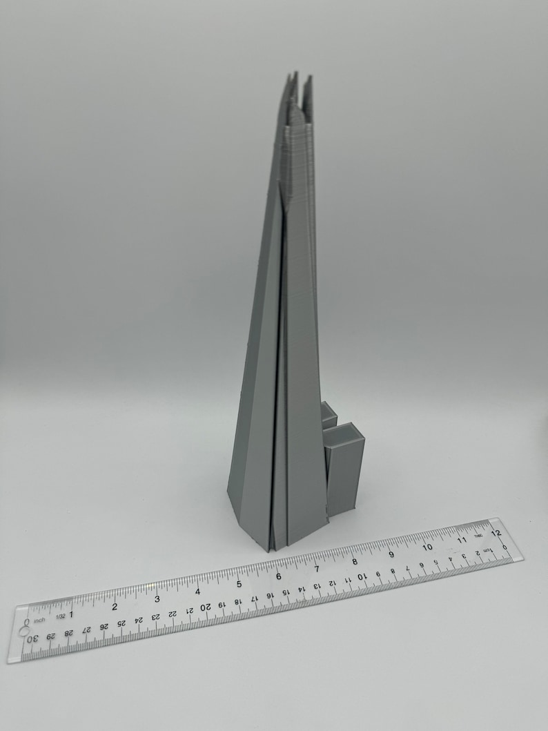 May include: A gray 3D printed model of the Shard, a skyscraper in London, England. The model is about 6 inches tall and is sitting on a white surface.