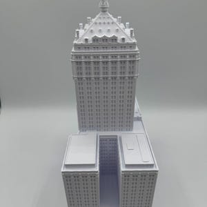 Puede incluir: Maqueta arquitect&oacute;nica blanca de un rascacielos. El modelo presenta una alta torre central con un techo y una aguja detallados, flanqueada por estructuras rectangulares inferiores. El edificio tiene numerosas ventanas y detalles arquitect&oacute;nicos.