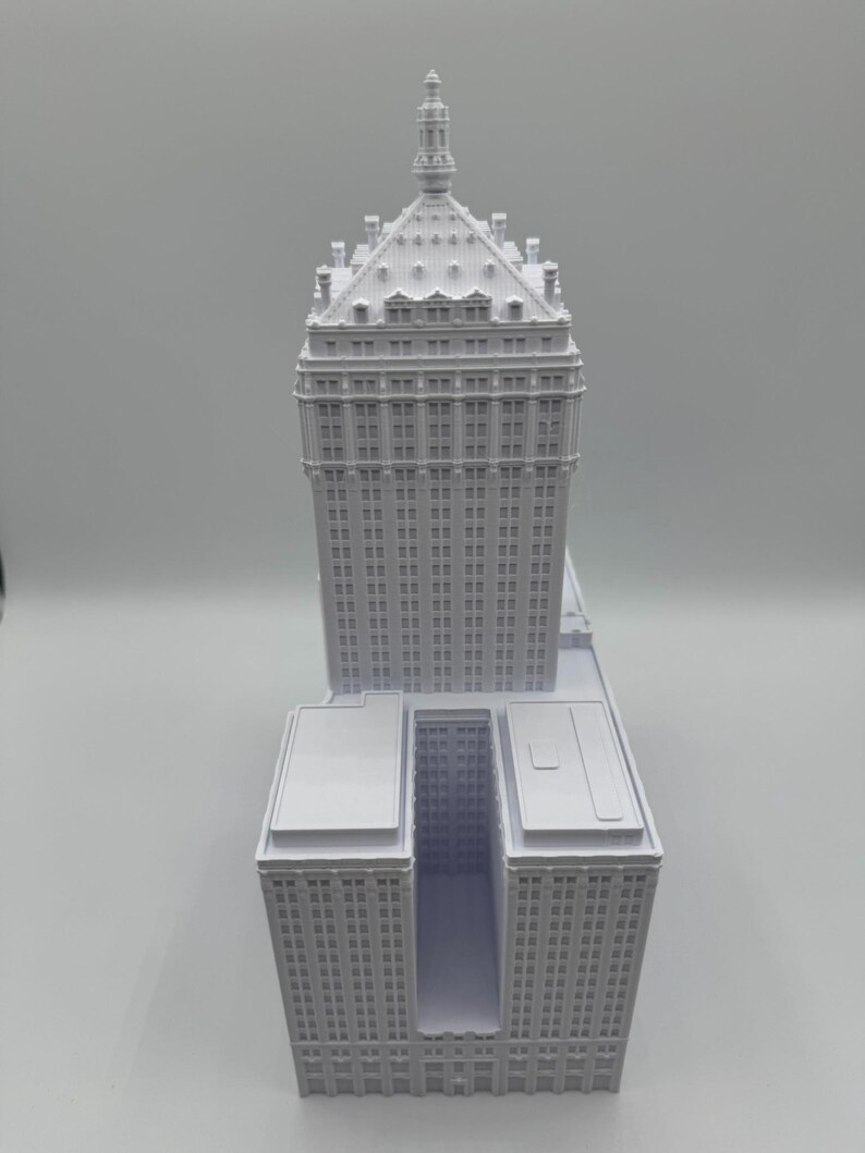 Puede incluir: Maqueta arquitect&oacute;nica blanca de un rascacielos. El modelo presenta una alta torre central con un techo y una aguja detallados, flanqueada por estructuras rectangulares inferiores. El edificio tiene numerosas ventanas y detalles arquitect&oacute;nicos.