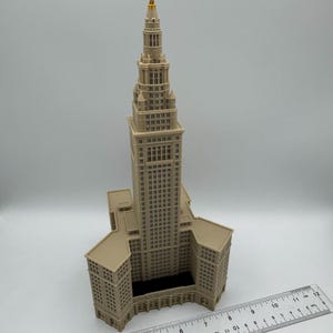 May include: A beige architectural model of a skyscraper with a gold-colored spire. The model has detailed windows and architectural features. A ruler is placed next to the model, showing measurements in inches.