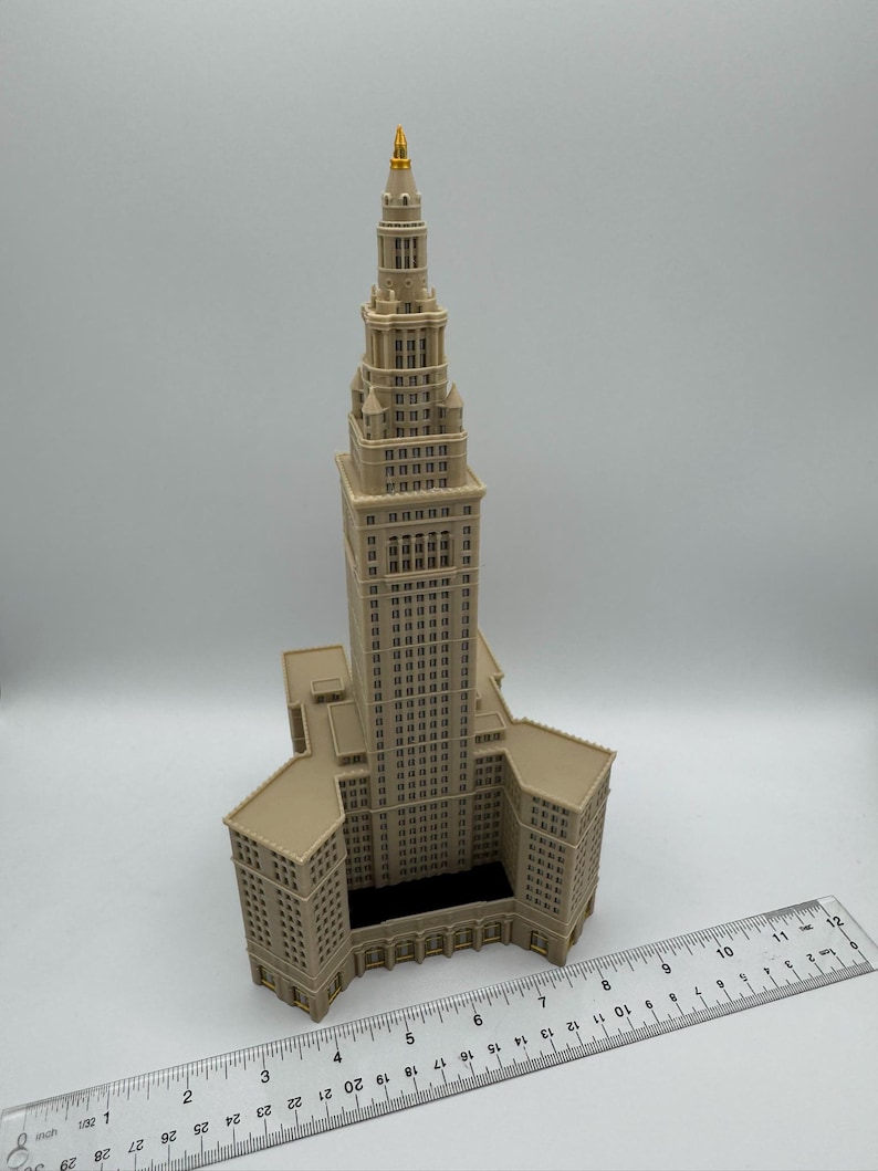 May include: A beige architectural model of a skyscraper with a gold-colored spire. The model has detailed windows and architectural features. A ruler is placed next to the model, showing measurements in inches.
