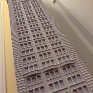 Op de afbeelding: Een wit 3D-geprint model van een hoog gebouw met veel ramen en decoratieve details. Het gebouw heeft een klok op de bovenkant.
