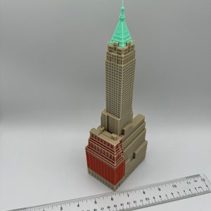 May include: A 3D printed model of a skyscraper with a green roof and a red brick facade. The model is approximately 15 cm tall and is sitting on a white surface.
