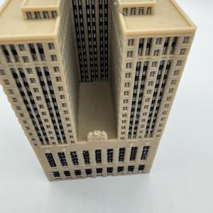 May include: A beige architectural model of a skyscraper with multiple stories and windows. The building has a rectangular shape with a central recessed area. The model is made of a light-colored material and has a detailed facade.