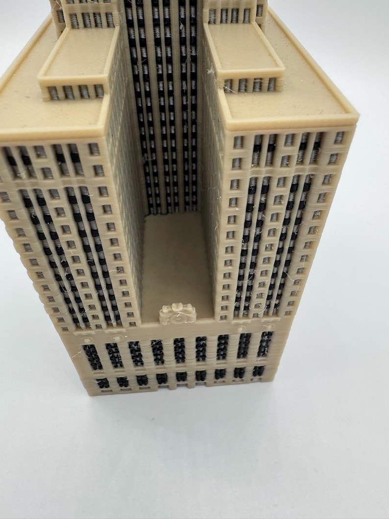 May include: A beige architectural model of a skyscraper with multiple stories and windows. The building has a rectangular shape with a central recessed area. The model is made of a light-colored material and has a detailed facade.