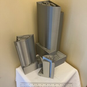 May include: Four 3D printed models of buildings, two tall and two shorter, in a light gray color. The models are arranged on a white tablecloth with a ruler in the foreground.