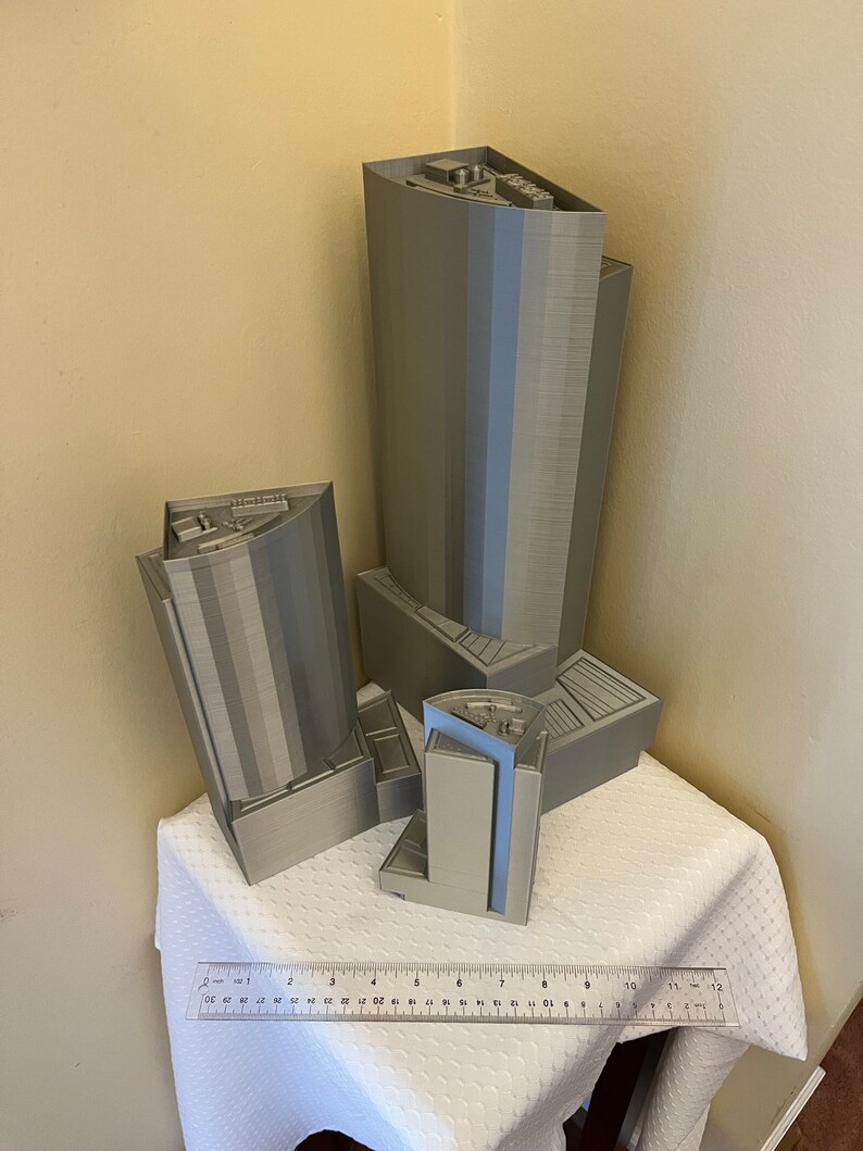 May include: Four 3D printed models of buildings, two tall and two shorter, in a light gray color. The models are arranged on a white tablecloth with a ruler in the foreground.