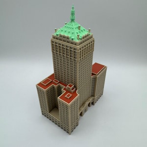 May include: A detailed miniature model of a skyscraper. The building features a tan facade, a green roof, and red accents. The model is a replica of a classic architectural design.