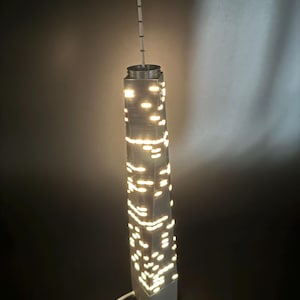 Könnte beinhalten: Ein weißes und graues beleuchtetes Modell eines Wolkenkratzers mit beleuchteten Fenstern. Das Modell hat eine hohe Antenne und einen weißen Sockel. Die Lichter sind hell vor dem dunklen Hintergrund. Ein weißes Kabel erstreckt sich von der Basis.