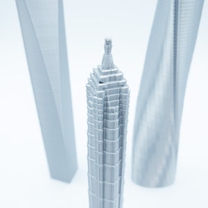 May include: A silver 3D printed model of a skyscraper with a pointed top. The model is surrounded by two other silver 3D printed skyscrapers.