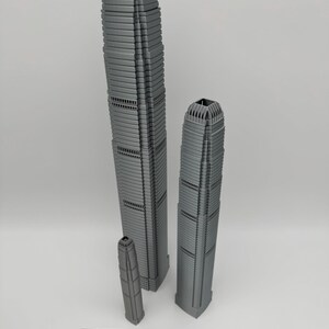 May include: Three gray miniature skyscraper models of varying heights. The tallest model is approximately 12 inches tall, the next is about 9 inches, and the smallest is around 4 inches. A wooden ruler is at the bottom.