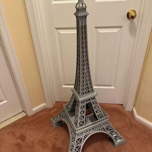 May include: A gray Eiffel Tower model, standing tall with intricate lattice detailing. The model is a replica of the iconic Parisian landmark, showcasing its architectural design. The tower is made of a light gray material and stands on a brown carpet.