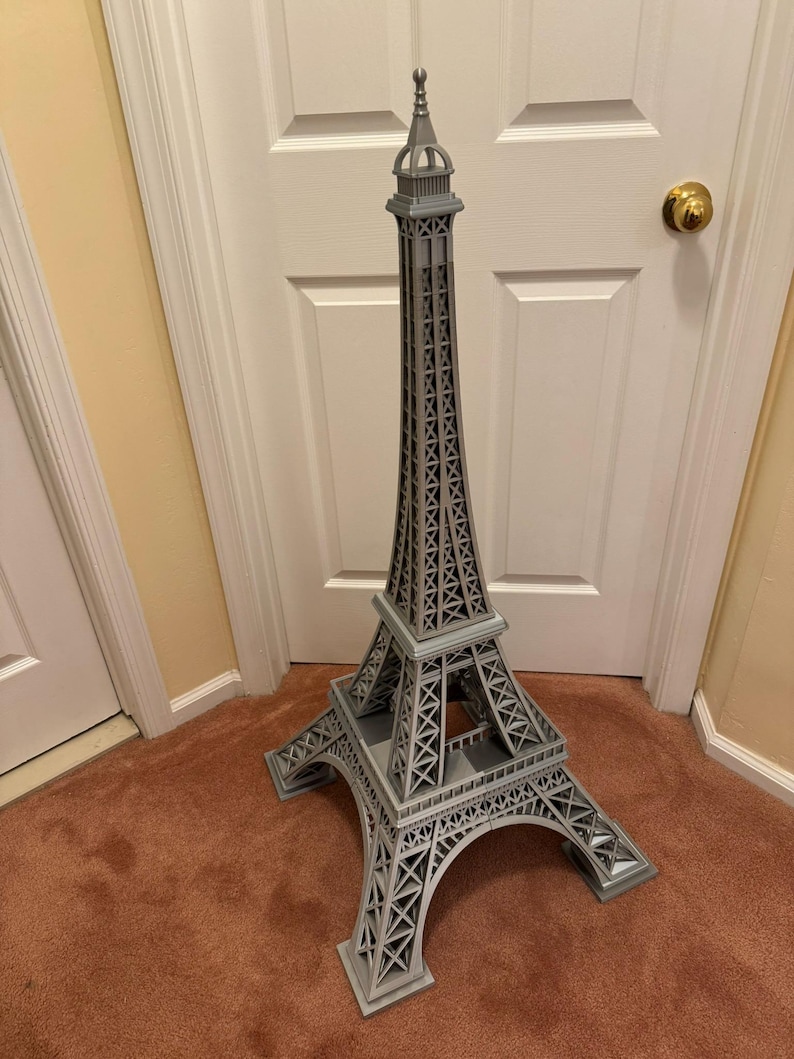 May include: A gray Eiffel Tower model, standing tall with intricate lattice detailing. The model is a replica of the iconic Parisian landmark, showcasing its architectural design. The tower is made of a light gray material and stands on a brown carpet.