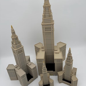 May include: A collection of beige miniature skyscraper models with gold-colored spires. The buildings vary in size and are arranged on a white surface. A ruler is placed at the bottom of the image.