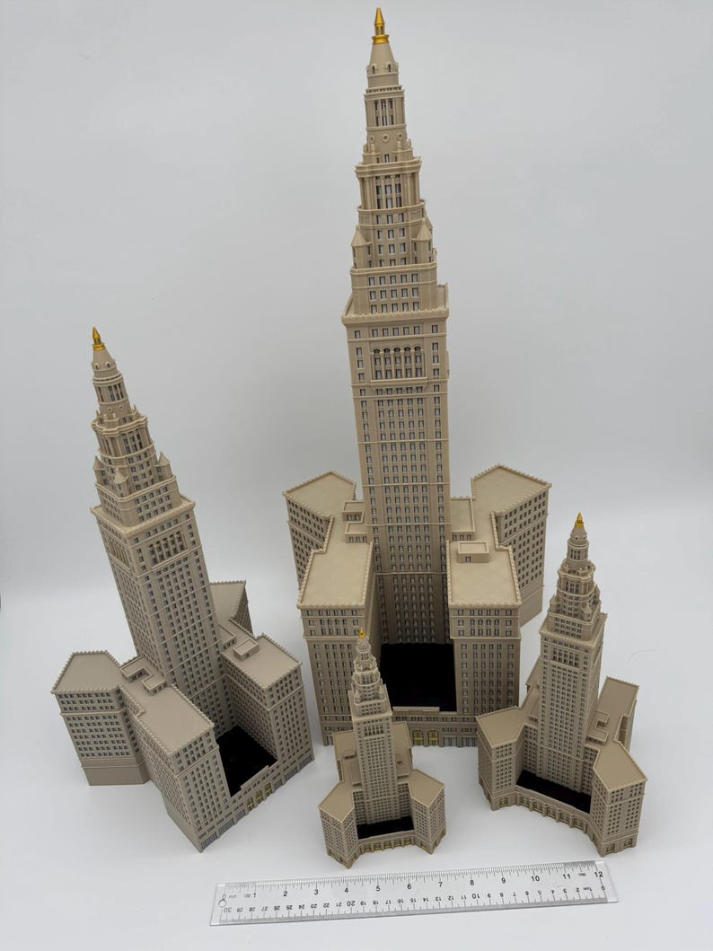 May include: A collection of beige miniature skyscraper models with gold-colored spires. The buildings vary in size and are arranged on a white surface. A ruler is placed at the bottom of the image.