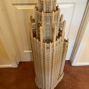 May include: A detailed 3D printed model of a tall, ornate building with a pointed roof and many windows. The model is made of a light brown plastic and is standing on a brown carpet.