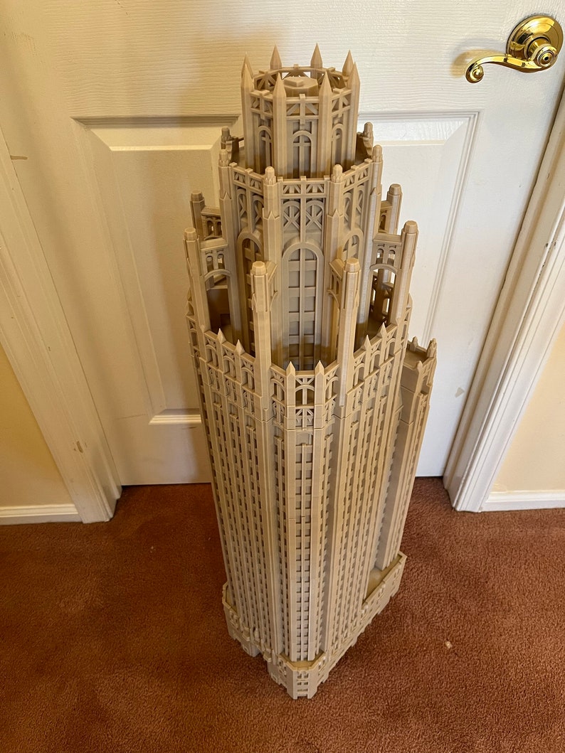 May include: A detailed 3D printed model of a tall, ornate building with a pointed roof and many windows. The model is made of a light brown plastic and is standing on a brown carpet.