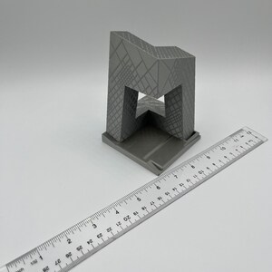 Peut inclure: Un mod&egrave;le d'immeuble imprim&eacute; en 3D de couleur grise avec un design g&eacute;om&eacute;trique. Le mod&egrave;le mesure environ 18 cm de haut et est pos&eacute; sur une surface blanche.