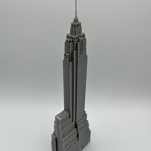 K&ouml;nnte beinhalten: Ein graues Miniaturmodell des Empire State Buildings. Das Modell ist sehr detailliert und zeigt das ikonische Art-Deco-Design des Geb&auml;udes, einschlie&szlig;lich seiner gestaffelten Struktur und Antenne. Das Geb&auml;ude besteht aus einem festen Material und steht auf einer wei&szlig;en Oberfl&auml;che.