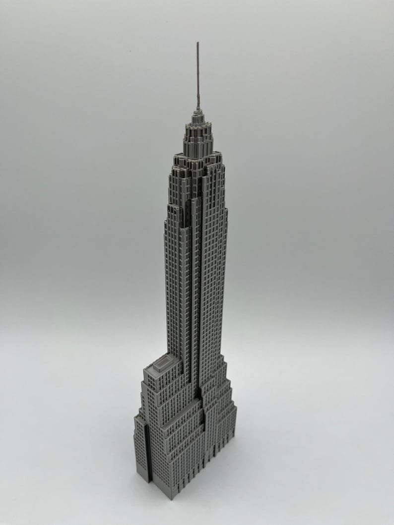 K&ouml;nnte beinhalten: Ein graues Miniaturmodell des Empire State Buildings. Das Modell ist sehr detailliert und zeigt das ikonische Art-Deco-Design des Geb&auml;udes, einschlie&szlig;lich seiner gestaffelten Struktur und Antenne. Das Geb&auml;ude besteht aus einem festen Material und steht auf einer wei&szlig;en Oberfl&auml;che.