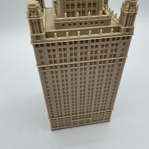 May include: A beige architectural model of a tall building with intricate details. The model features multiple levels, windows, and decorative elements, showcasing a classic skyscraper design. The building has a rectangular base and a central tower.