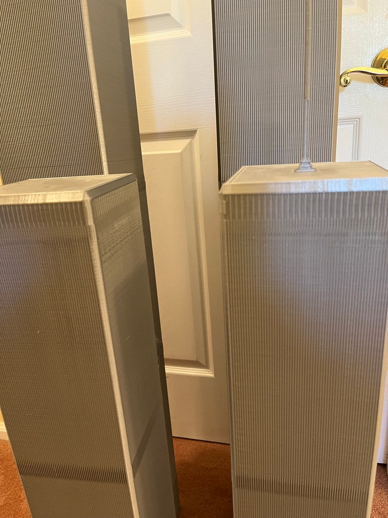 Pu&ograve; includere: Modelli in miniatura delle Torri Gemelle, di colore grigio chiaro, con dettagli a coste verticali. I modelli sono posizionati davanti a una porta bianca e un tappeto marrone.