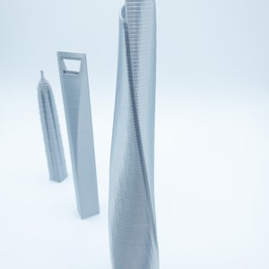 May include: Three silver 3D printed models of skyscrapers. The tallest model is a twisted tower with a smooth, curved surface. The other two models are more traditional, with straight lines and a rectangular shape.