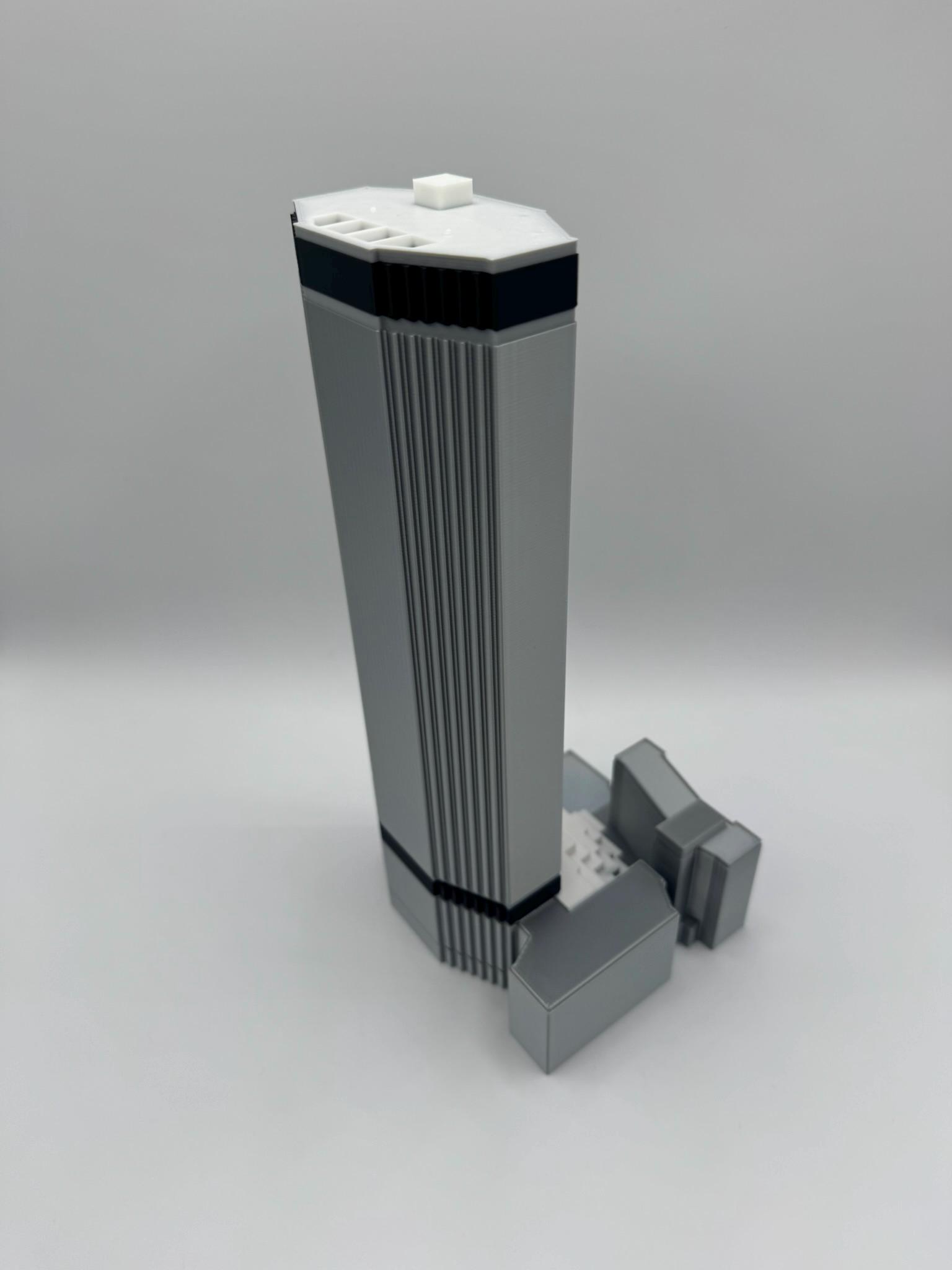 May include: A gray architectural model of a skyscraper with a rectangular base and a tall, vertical structure. The model has a white top with a small square element and black accents. The building has vertical lines and a smaller structure attached.