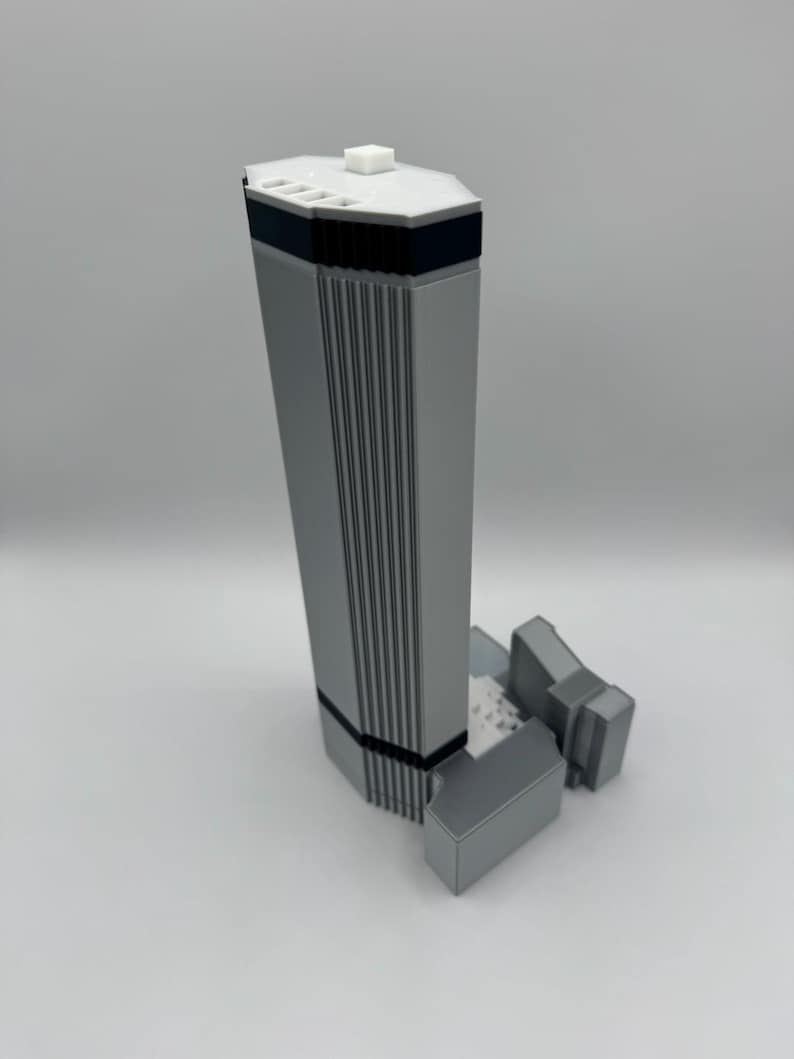 May include: A gray architectural model of a skyscraper with a rectangular base and a tall, vertical structure. The model has a white top with a small square element and black accents. The building has vertical lines and a smaller structure attached.