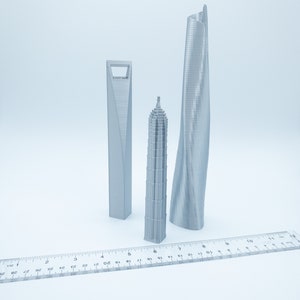 May include: Three silver 3D printed models of skyscrapers, a tall, thin, curved building, a shorter, wider building with a square base, and a tall, thin, pointed building. A ruler is in the foreground.