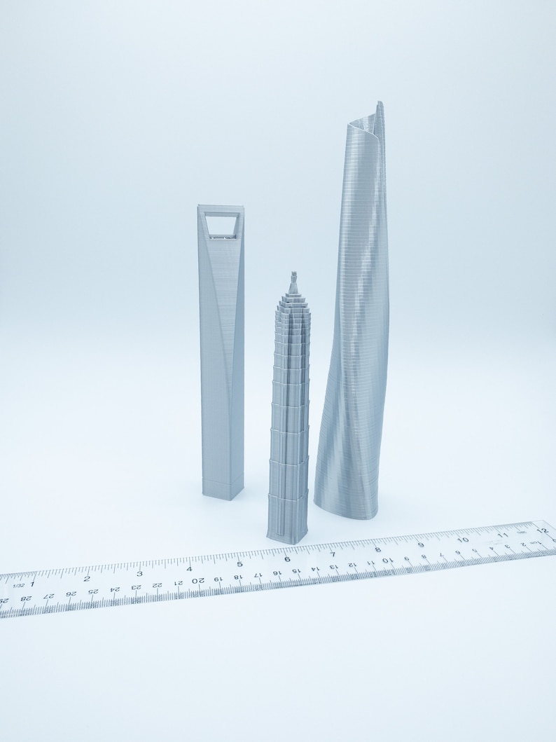 May include: Three silver 3D printed models of skyscrapers, a tall, thin, curved building, a shorter, wider building with a square base, and a tall, thin, pointed building. A ruler is in the foreground.