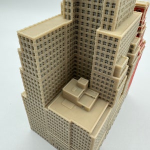 May include: A detailed 3D printed model of a tall beige building with multiple windows and a flat roof. The building has a slightly angled roof and a small rectangular structure on the top.