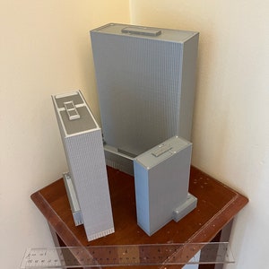 May include: Three gray 3D printed model buildings of varying sizes on a wooden table with a ruler.