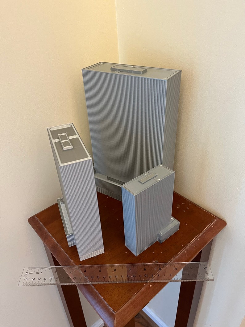 May include: Three gray 3D printed model buildings of varying sizes on a wooden table with a ruler.