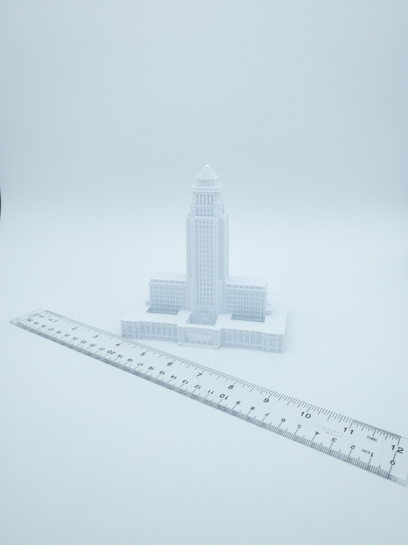 May include: A white 3D printed model of a city hall building. The model is about 6 inches tall and is sitting on a white ruler.