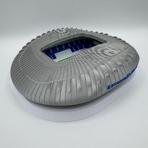 May include: A gray stadium model with a ridged roof and a white base. The interior shows a green field and blue seating. The text "ESTADIO" is visible on the side. A detailed miniature of a sports arena.